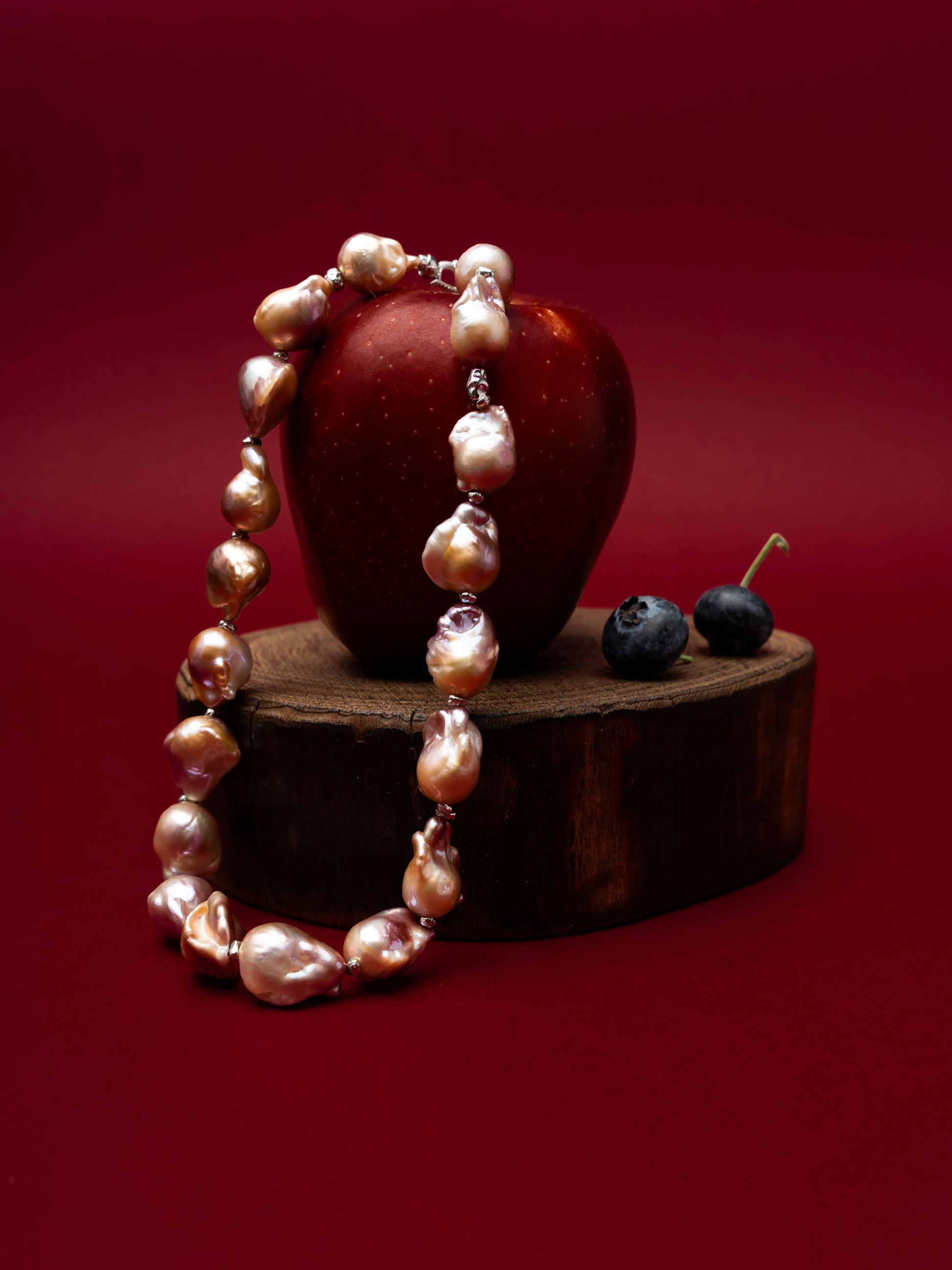 Pearl necklace draped over a red apple on a wooden stand with blueberries on a red background