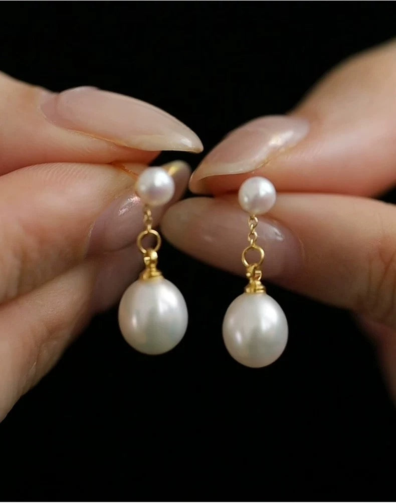 Pearl earrings with gold hooks held between fingers against a black background