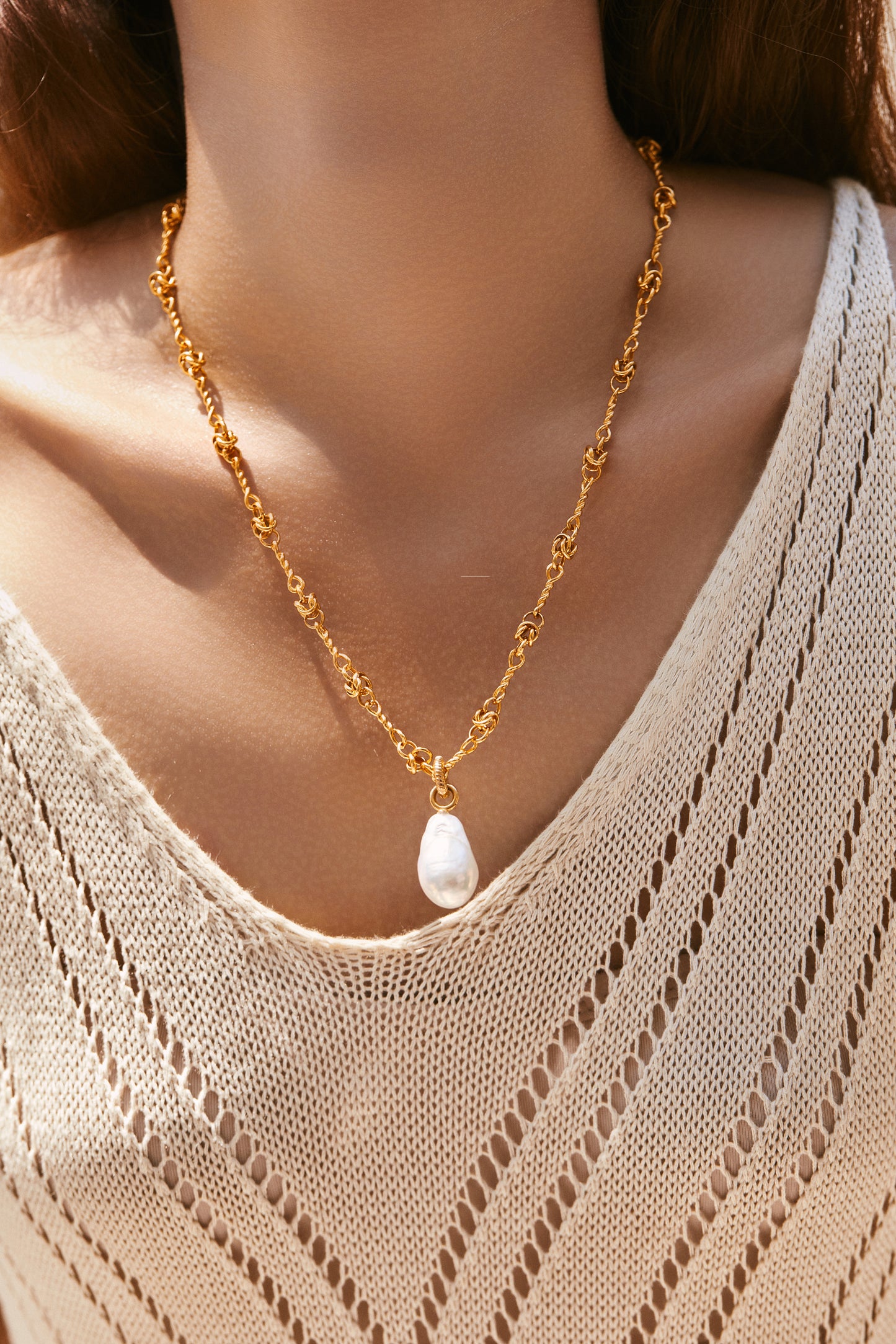 Gold chain necklace with a baroque pearl pendant worn by a person in a beige sweater.