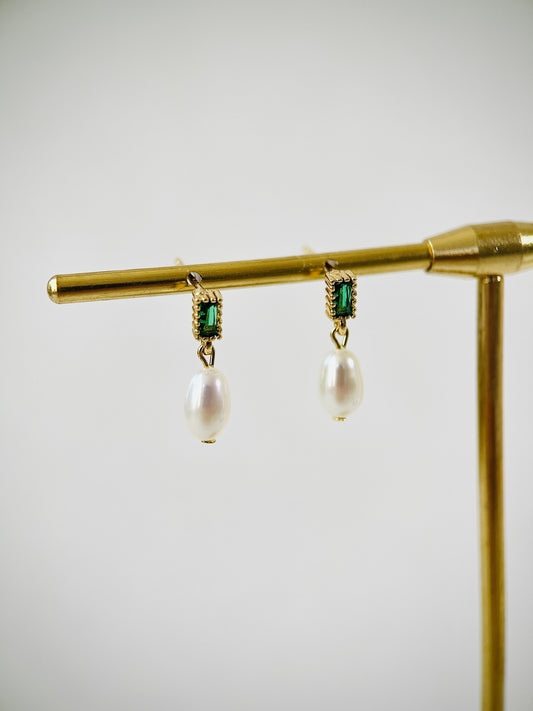 Pearl earrings with green gemstones on a gold stand against a white background